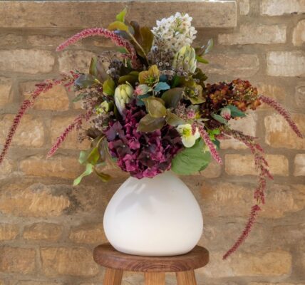A lush arrangement of dark purple hydrangeas, white flowers, and trailing pink amaranthus in a round white ceramic vase, sitting on a wooden stool against a rustic stone wall.