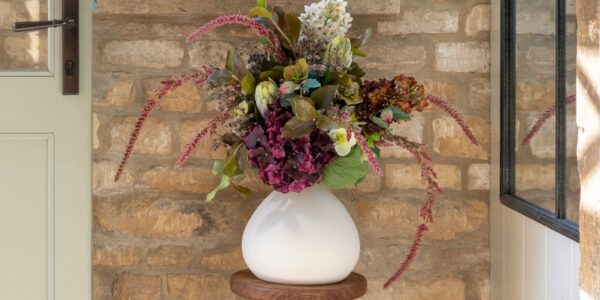 A lush arrangement of dark purple hydrangeas, white flowers, and trailing pink amaranthus in a round white ceramic vase, sitting on a wooden stool against a rustic stone wall.