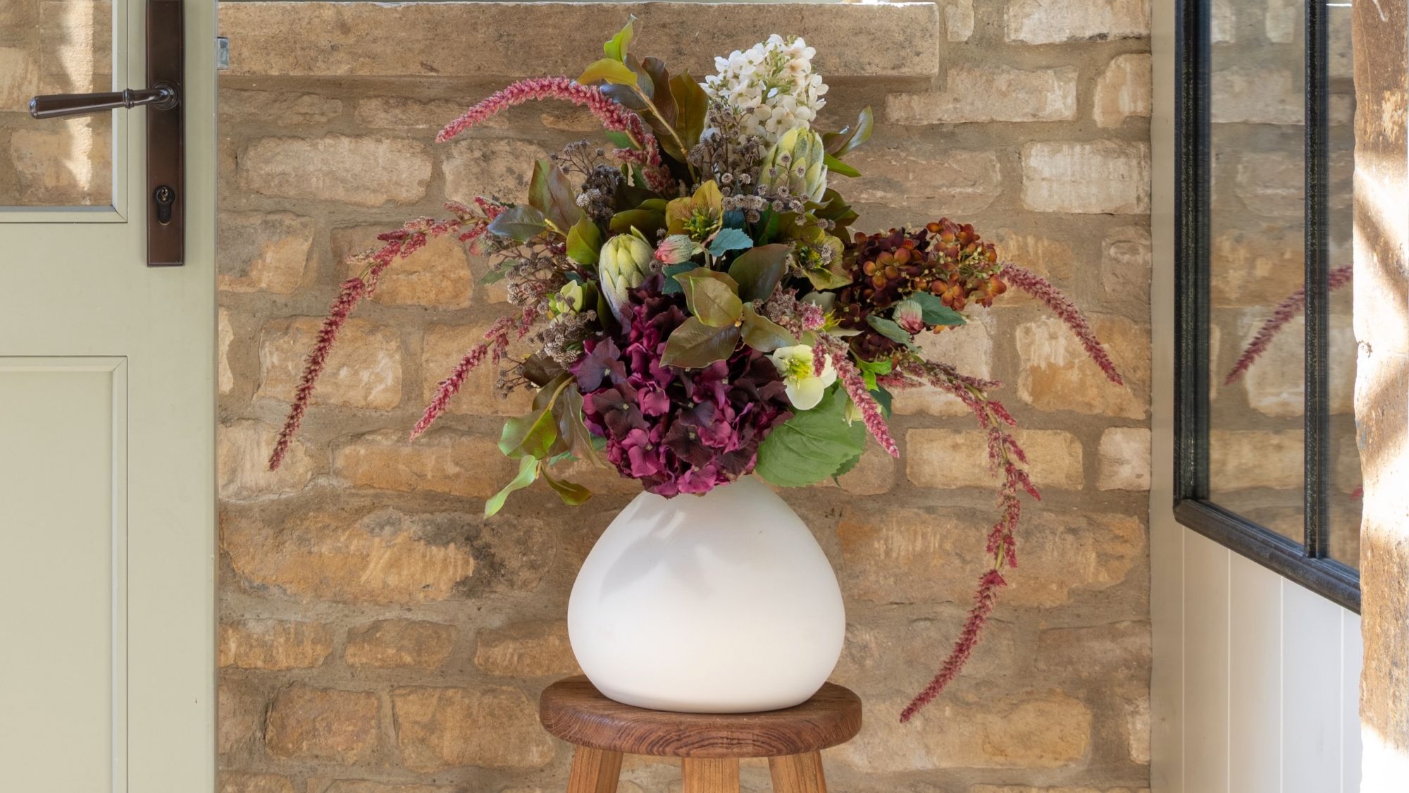 A lush arrangement of dark purple hydrangeas, white flowers, and trailing pink amaranthus in a round white ceramic vase, sitting on a wooden stool against a rustic stone wall.