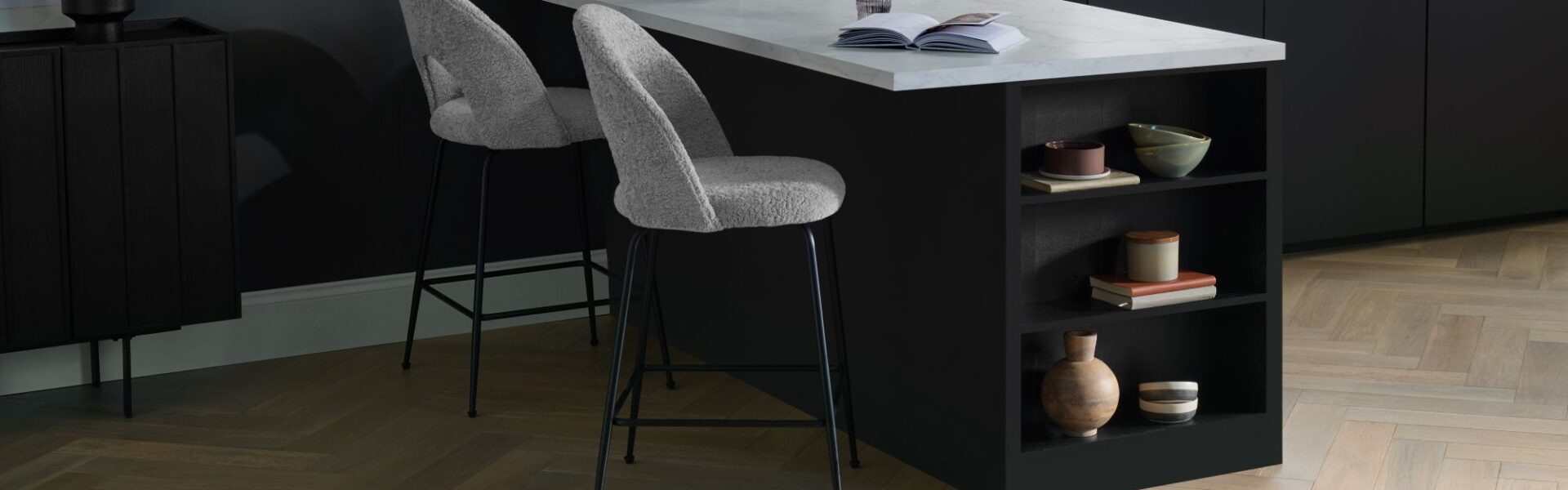 Modern kitchen featuring light oak herringbone engineered wood flooring, a black kitchen island with white marble countertop, and grey bouclé bar stools.