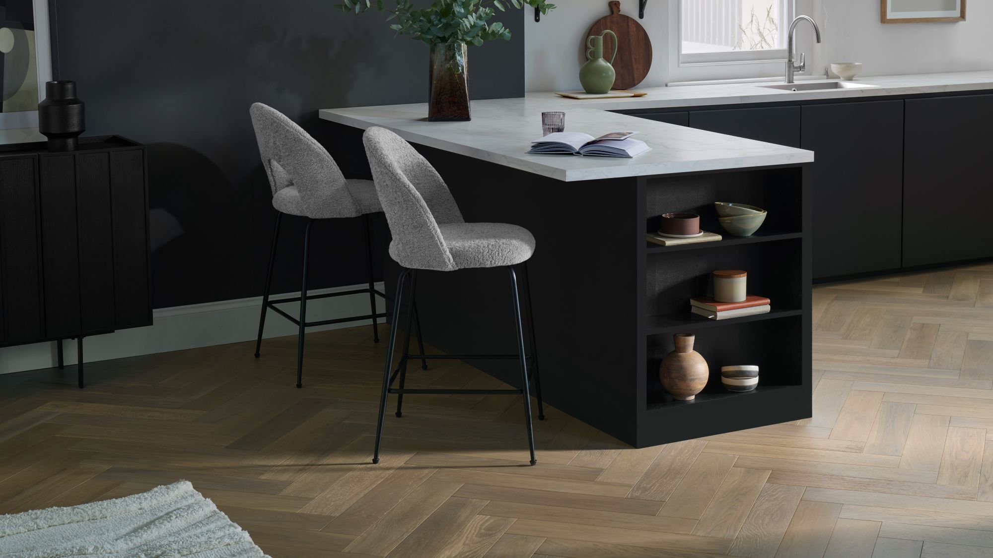 Modern kitchen featuring light oak herringbone engineered wood flooring, a black kitchen island with white marble countertop, and grey bouclé bar stools.