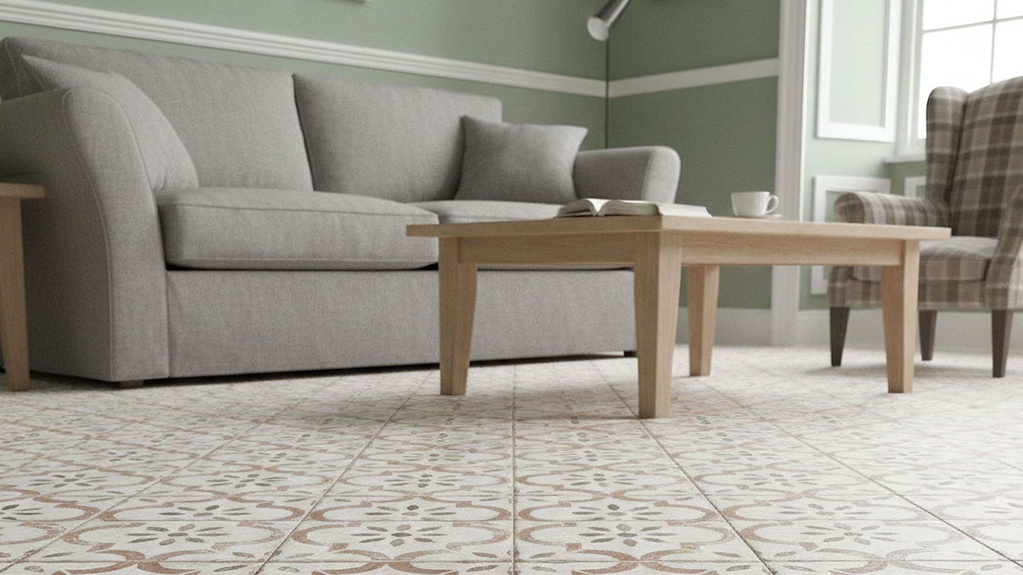 Living room with light patterned Salcombe Cotto floor tiles, featuring a grey couch, a light wood coffee table with an open book, sage green walls, and a plaid armchair by the window.