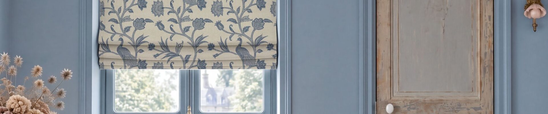 Escape to the Chateau L'Oiseau du Chateau Roman blind in blue featuring a vintage botanical bird and floral pattern, hung over a window in a blue room with a distressed wooden door