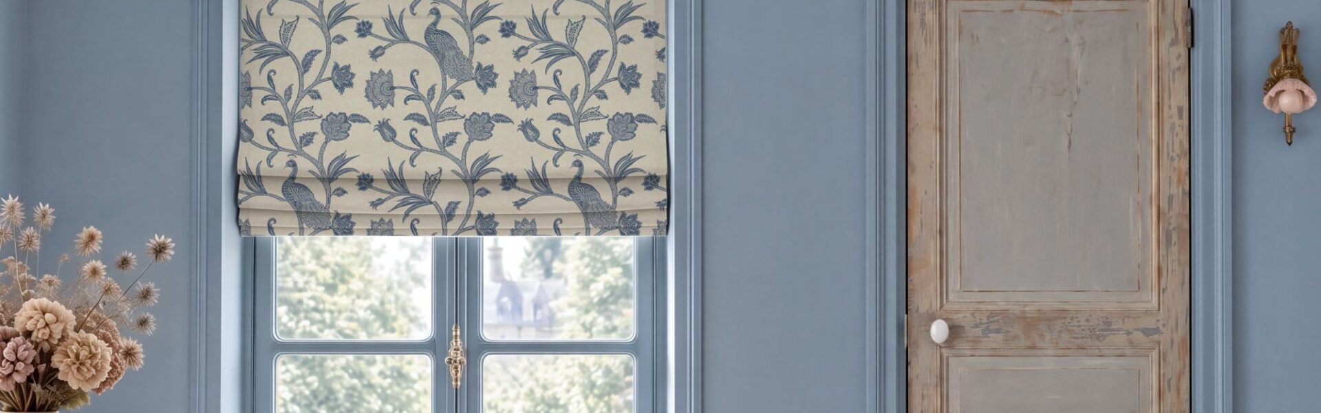 Escape to the Chateau L'Oiseau du Chateau Roman blind in blue featuring a vintage botanical bird and floral pattern, hung over a window in a blue room with a distressed wooden door