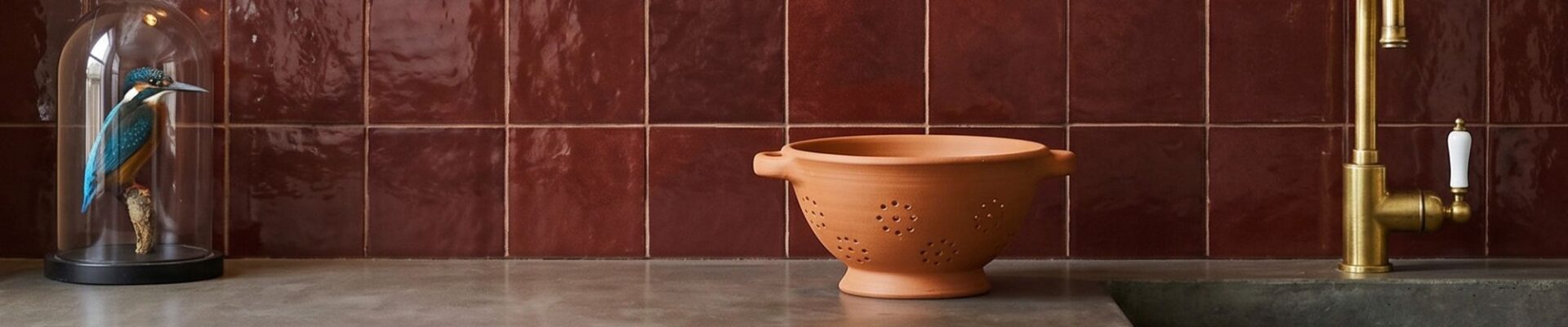 Glossy ruby-red square tiles backsplash in a kitchen with a concrete countertop, terracotta colander, and brass tap.