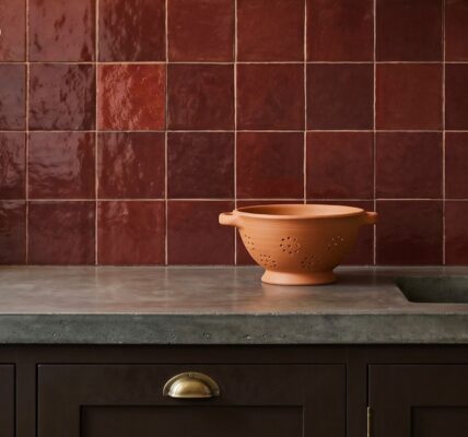 Glossy ruby-red square tiles backsplash in a kitchen with a concrete countertop, terracotta colander, and brass tap.
