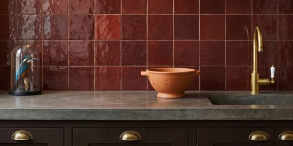 Glossy ruby-red square tiles backsplash in a kitchen with a concrete countertop, terracotta colander, and brass tap.
