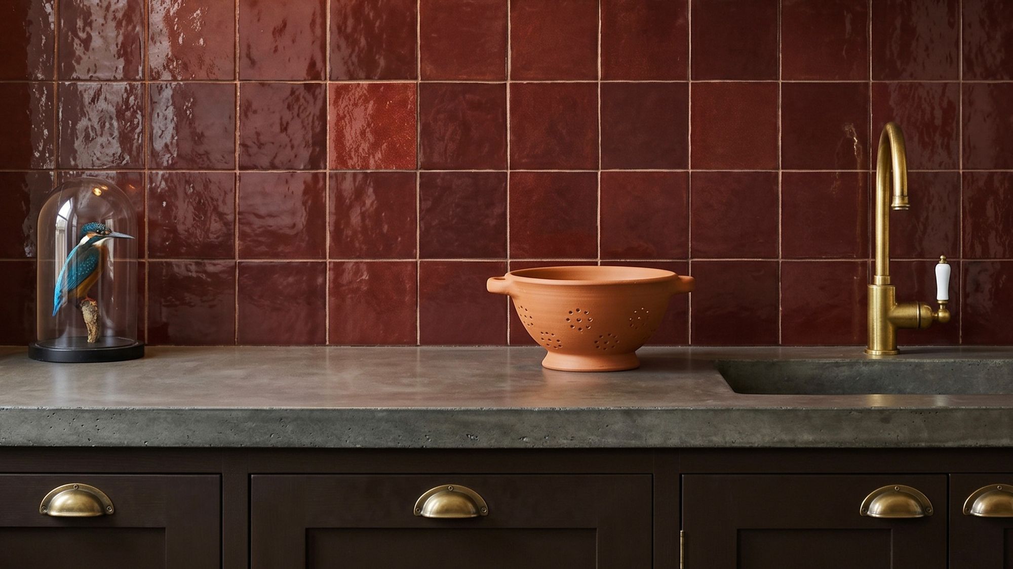 Glossy ruby-red square tiles backsplash in a kitchen with a concrete countertop, terracotta colander, and brass tap.