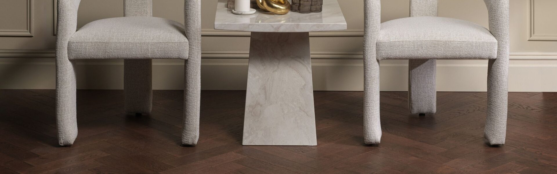 Two light grey upholstered accent chairs set against wainscoted walls, featuring a small marble side table between them on dark brown oak herringbone parquet flooring.
