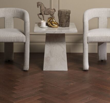 Two light grey upholstered accent chairs set against wainscoted walls, featuring a small marble side table between them on dark brown oak herringbone parquet flooring.