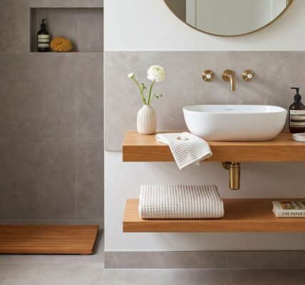 A modern bathroom featuring double floating wooden shelves, a white vessel sink, brushed brass wall-mounted fixtures, and grey wall and floor tiles.