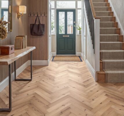 Bright and stylish hallway featuring burnished oak herringbone LVT flooring, a live-edge console table with dried florals, slatted wood accent walls, and a staircase with a textured runner leading to a dark green front door