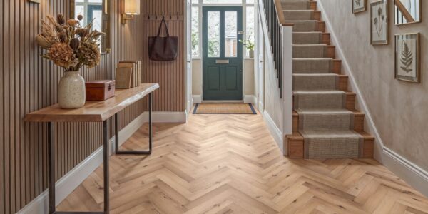 Bright and stylish hallway featuring burnished oak herringbone LVT flooring, a live-edge console table with dried florals, slatted wood accent walls, and a staircase with a textured runner leading to a dark green front door